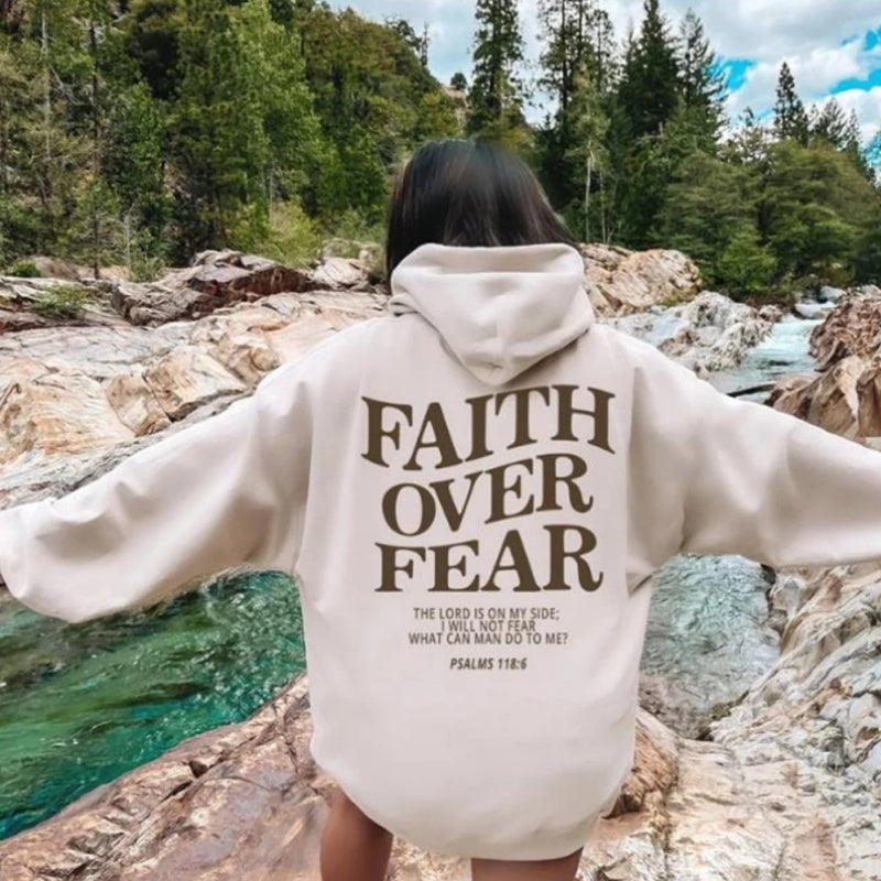 A person stands on rocks by a river and wears the Faith Over Fear Christian Hoodie, a trendy unisex sweatshirt featuring a bold Bible verse, with a forest background highlighting this inspirational moment.