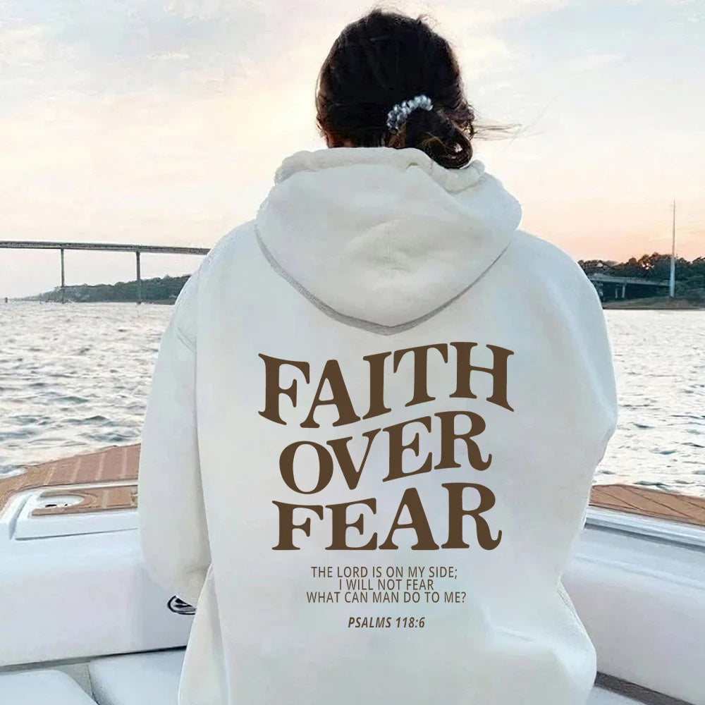Someone wearing the Faith Over Fear Christian Hoodie sits on a boat facing away. The hoodie features "FAITH OVER FEAR. The Lord is on my side; I will not fear. What can man do to me? PSALMS 118:6." A bridge and water are in the background.