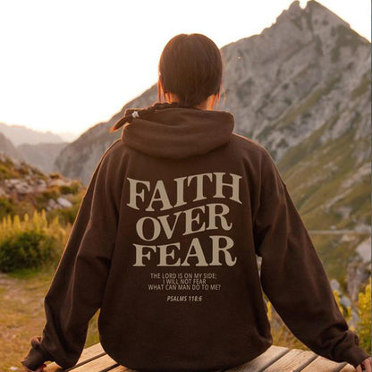 A person sits outdoors at sunset facing mountains, wearing the Faith Over Fear Christian Hoodie with bold back text: “FAITH OVER FEAR. The Lord is on my side; I will not fear. What can man do to me? Psalms 118:6.”.