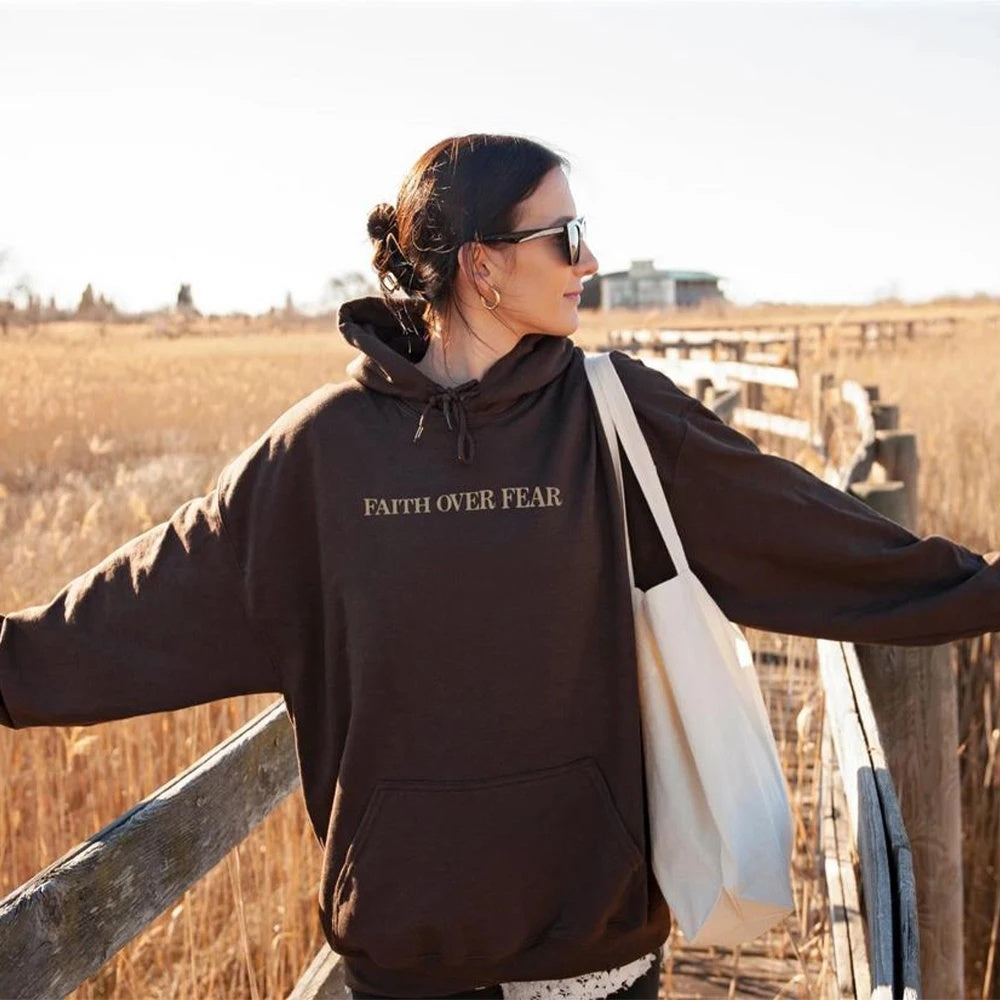 A woman in sunglasses wears a dark "Faith Over Fear" Christian Hoodie (Trendy Bible Verse Unisex Sweatshirt) as she walks on a wooden path through tall grass, carrying a white tote bag.